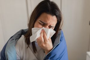 Mulher cobrindo-se com um cobertor enquanto segura um lenço, aparentando estar resfriada ou lidando com sintomas de gripe, como espirros e congestão nasal.