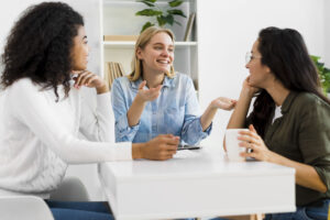 tres mulheres jovens conversam e sorriem ao redor de uma mesa 