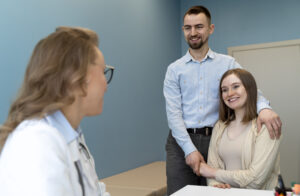 A imagem mostra uma consulta médica com três pessoas em um ambiente clínico. À esquerda, uma médica, com jaleco branco e óculos, parece estar conversando ou dando orientações. Ao centro e à direita, um casal, provavelmente paciente e acompanhante, parece estar interagindo com a médica. A mulher está sorrindo e parece relaxada, enquanto o homem, também sorrindo, coloca a mão sobre o ombro dela, transmitindo apoio. Eles estão em pé, e a médica está sentada. O ambiente tem paredes azuis e uma mesa branca com alguns objetos, sugerindo um consultório médico. A cena transmite uma atmosfera amigável e de suporte durante a consulta.