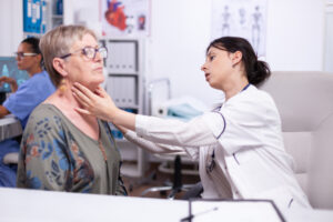 médica jovem examinando o pescoço de uma paciente idosa em um consultório. A médica está de jaleco branco e a paciente, de cabelos grisalhos e óculos, veste uma blusa estampada. A médica toca o pescoço da paciente verificando desconforto na garganta. Ao fundo, há elementos de um ambiente clínico, como estantes com pastas e pôsteres anatômicos.