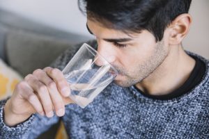 A imagem mostra um homem bebendo água de um copo transparente. Ele segura o copo com a mão direita enquanto aproxima o rosto para tomar o líquido. O homem está usando um suéter cinza de malha e tem cabelo escuro e barba por fazer. Ele está sentado em um ambiente confortável, que parece ser uma sala de estar, devido à presença de um sofá e almofadas ao fundo. 