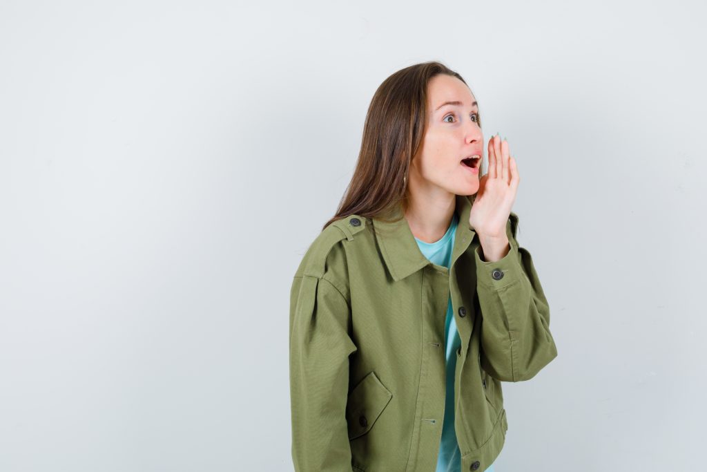 Mulher jovem usando jaqueta verde e camiseta azul, gesticulando com a mão em direção à boca como se estivesse chamando ou gritando, causa da cicatriz nas pregas vocais.