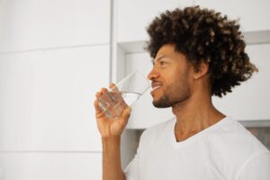 Homem de cabelo cacheado e barba, vestindo uma camiseta branca, segurando um copo de água e sorrindo enquanto bebe.
