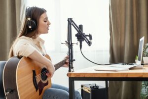 Mulher jovem com cabelo longo e liso, usando fones de ouvido e tocando um violão enquanto canta em um microfone profissional.